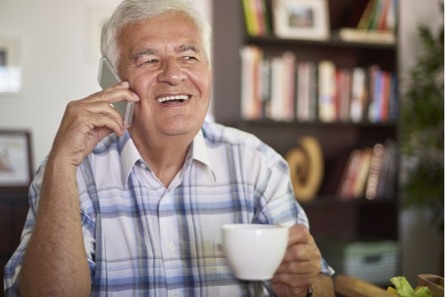äldre man pratar i telefon i vardagsrum med en kaffekopp i handen