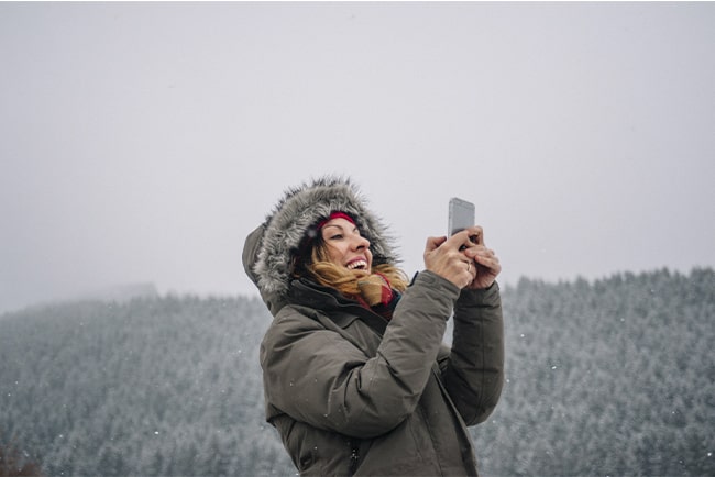 Kvinna håller upp mobil, med snötäckt skog i bakgrunden.